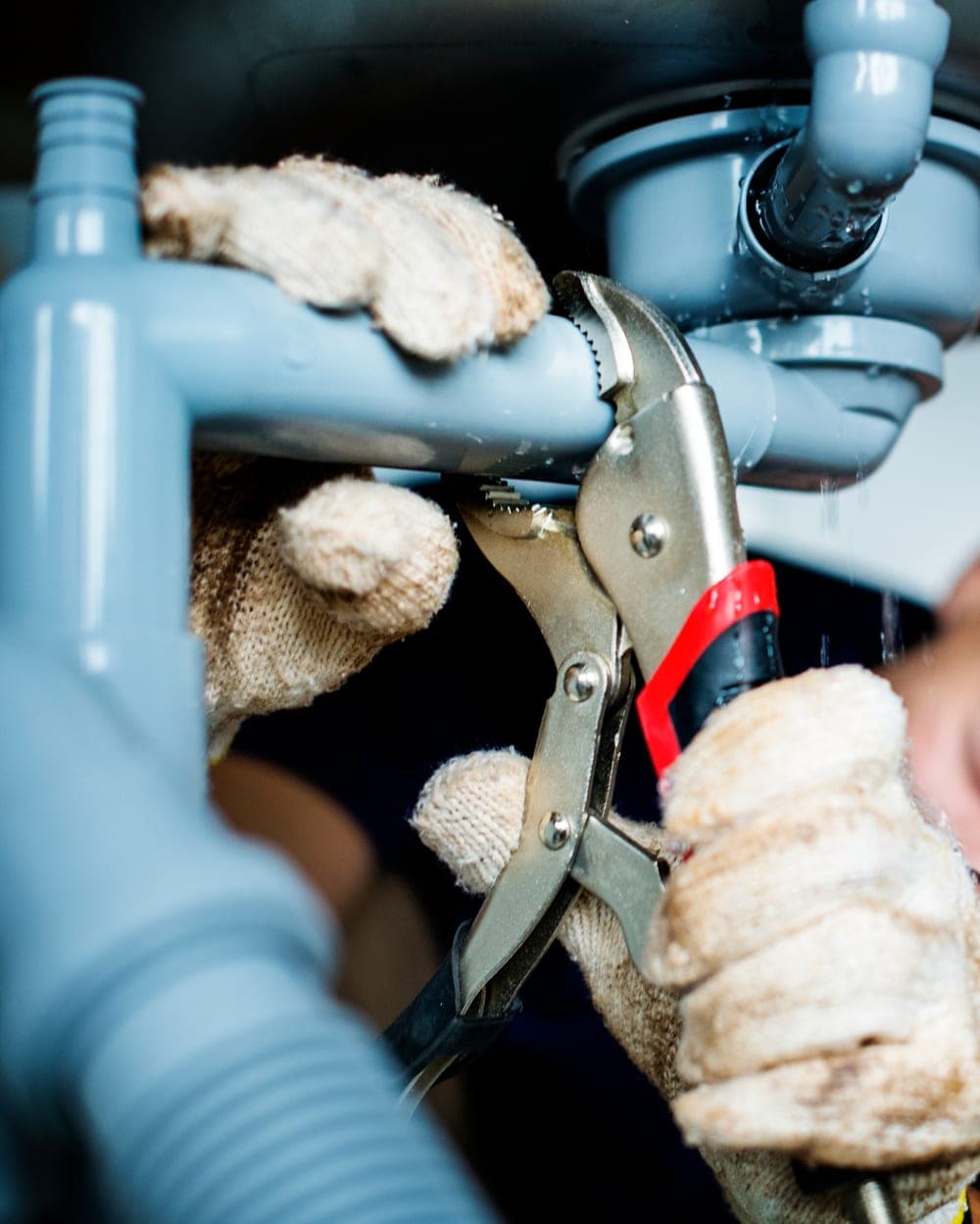 A plumber's gloved hands gripping a waste pipe with pliers under a sink
